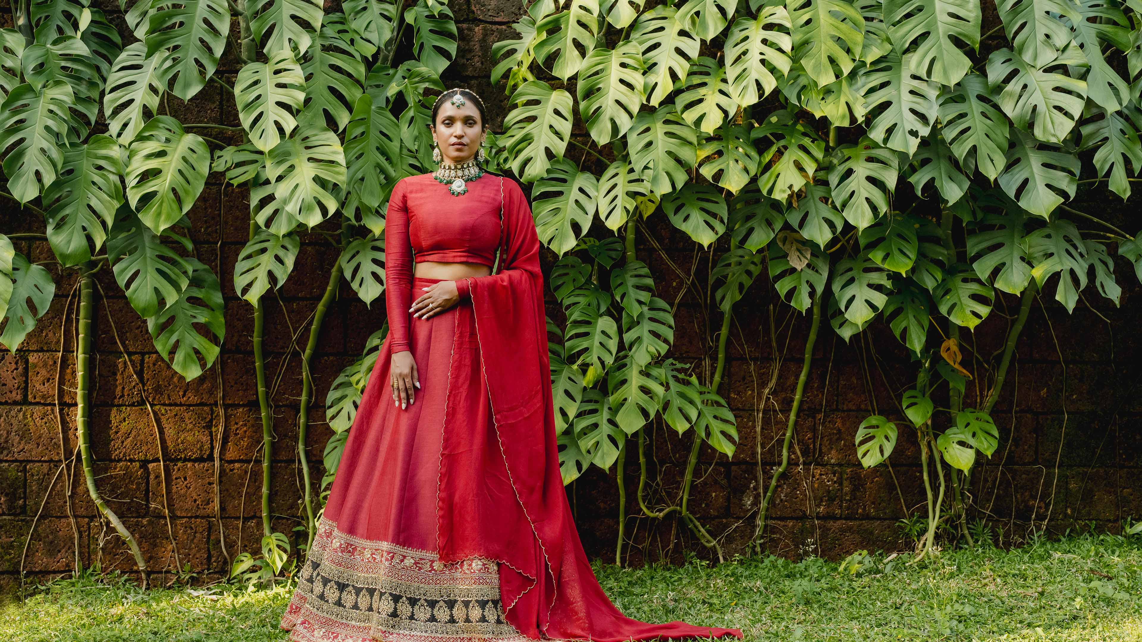 Woman in a red traditional outfit standing in front of a leafy green wall.